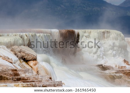 Hot mineral water cascading over stalactite formations with steam rising against the mountains at Yellowstone National Park.