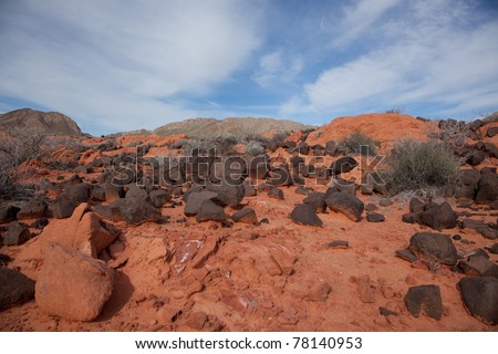 Randomly scattered black volcanic rocks sit on top of bright orange sandstone earth with a blue sky background in the Nevada desert near Northern Lake Mead.