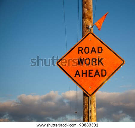 A scratched orange road work ahead sign with one tattered flag on top is bolted to a roadside power and telephone pole with a blue sky background.