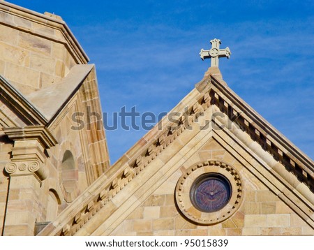 The steep roof lines of The Cathedral Basilica of St. Francis of Assisi with details of a cross and stained glass window,against a blue sky.