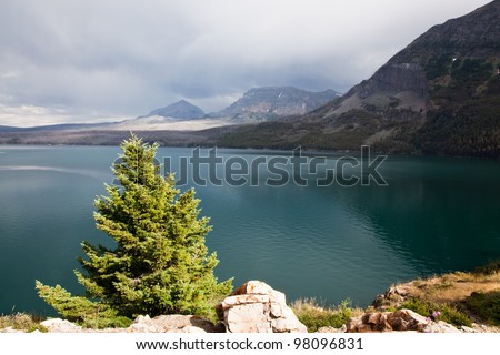A evergreen tree and rocky shoreline are lite by sunshine with Saint Mary Lake and surrounding mountains still in shadow from a passing storm in Glacier National Park, Montana.