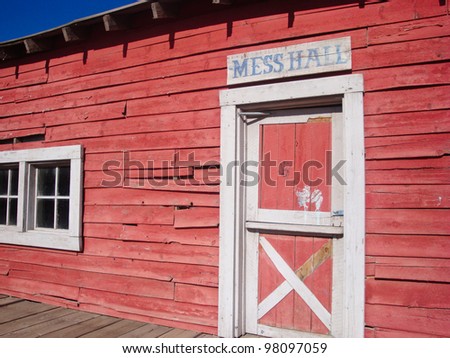 A mess hall sign above the white trimmed split door of an old western red barn building made from wood.