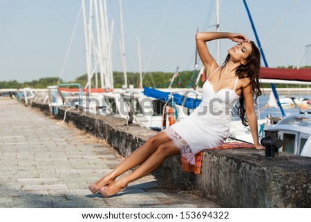 Beautiful woman in white dress sitting in the hot sun on the pier near the boats and yachts. Outdoors, lifestyle