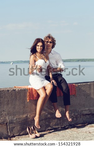 lovely beautiful couple sitting in the hot sun on the pier in summer. Young man and sensual brunette outdoor portrait in classic dress near the ocean. Outdoors, lifestyle.