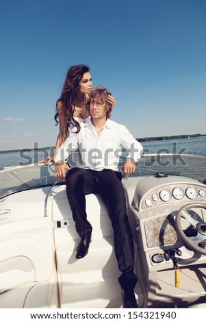 Sexy  beautiful couple posing on the luxury boat in open sea in summer. Young man and sensual brunette outdoor portrait in classic dress. Outdoors, lifestyle.