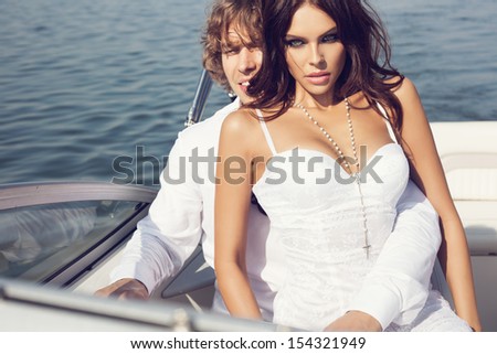 Sexy beautiful couple posing on the luxury boat in open sea in summer. Young man and sensual brunette outdoor portrait in classic dress. Outdoors, lifestyle.