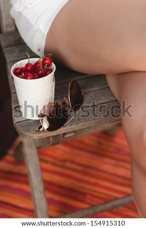A glass of cherry and sunglasses are on a chair near the feet of a young woman. Toned with tender summer color.