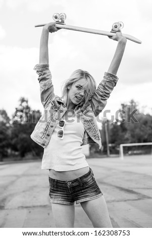A young woman is standing on a school playground and holding skateboard over her head. The girl in joyful feelings. Black and white. Outdoors, lifestyle.