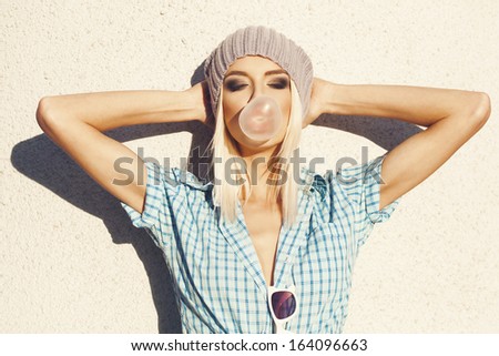 Sexy beautiful blonde in grey knit hat and blue shirt posing on wall background. Blow bubblegum. Outdoors, lifestyle.