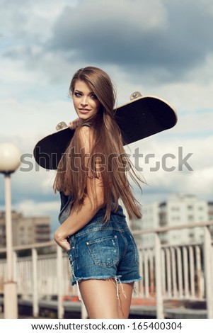 Young woman walking on street and holding skateboard behind her head. Pretty girl looking at camera. Outdoors, lifestyle.