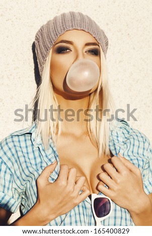 Trendy beautiful blond woman in grey knit hat and  blue shirt. Girl blowing bubblegum. Outdoors, lifestyle.