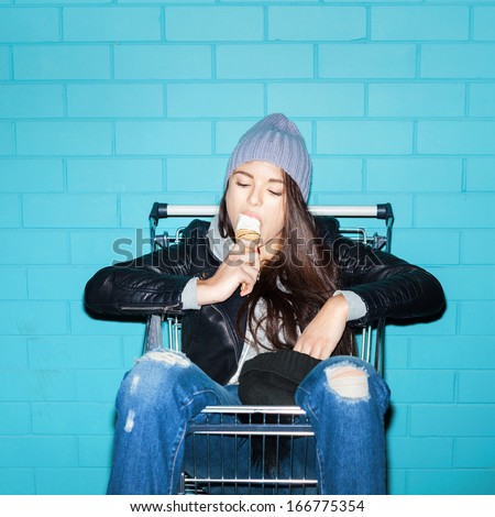 Young funky woman eating ice cream over blue brick wall. Naughty girl having fun in shopping cart. Indoors, lifestyle