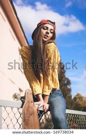 Beautiful young girl with longboard sitting on the fence in the afternoon, outdoor