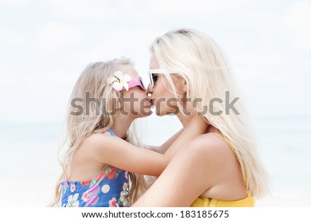 Young beautiful mother embracing and kissing her daughter sitting on a sea beach. Outdoors