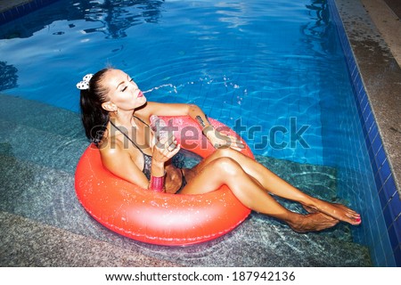 Sexual  woman floating in an pink inner tube in a swimming pool and drinking water. Outdoors, lifestyle
