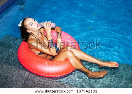 Funky young woman floating in an pink inner tube in a swimming pool and drinking water. Outdoors, lifestyle