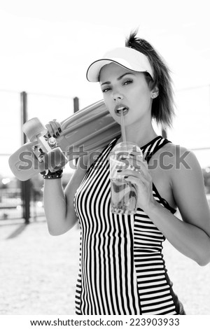 Sporty girl with skateboard drinking water against the sky. Black white portrait