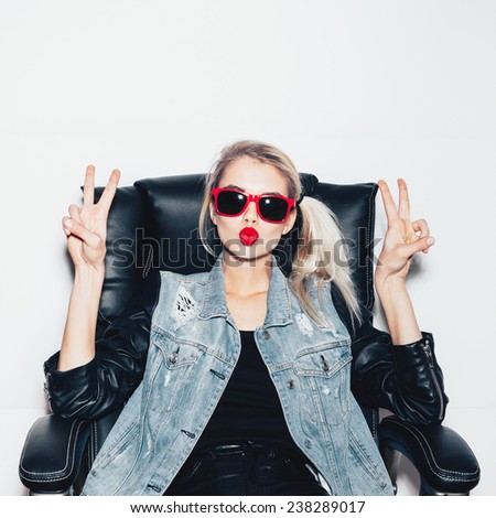 Young blonde woman in sunglasses sit on black office chair and showing victory by finger. Fashoin hipster girl.  White background, not isolated