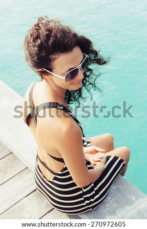Young pretty woman in sunglasses sitting alone on the pier near the sea and looking on the water and waiting for somebody in ocean