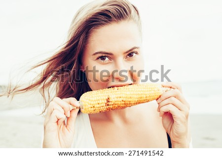 Outdoor summer style fashion portrait of young beautiful sensual woman posing on the beach on vacation. Girl holding corn. Snacking on the sea.