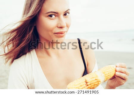 Outdoor summer style fashion portrait of young beautiful sensual woman posing on the beach on vacation. Girl holding corn and looking at camera. Snacking on the sea.