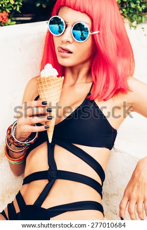 Woman with sunglasses in red wig and black bikini eating ice cream. Fashion stylish sexy girl. Outdoors lifestyle portrait