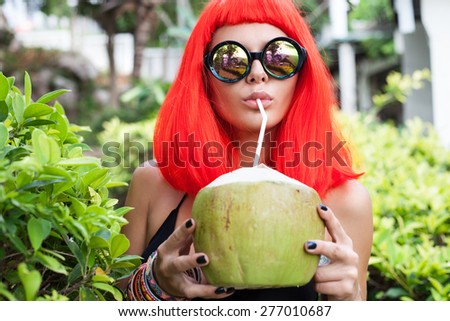 Woman with sunglasses in red wig and black bikini drinking coconut juice. Fashion stylish sexy girl. Outdoors lifestyle portrait