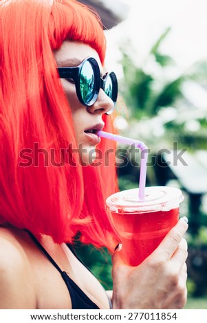 Woman with sunglasses in red wig and black bikini drinking cocktail. Outdoors lifestyle portrait