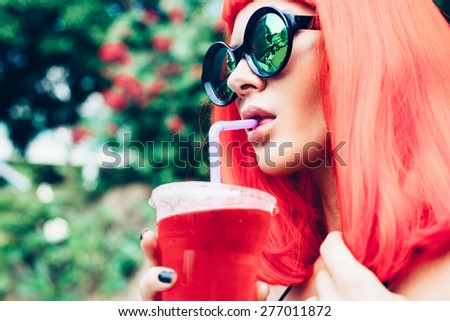 Woman with sunglasses in red wig and black bikini drinking cocktail. Outdoors lifestyle portrait