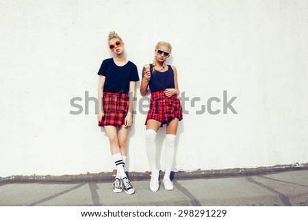 portrait of two pretty hipster blonde sisters  wearing plaid skirt and  black T-shirt. Girls smile, have fun against  urban white wall.