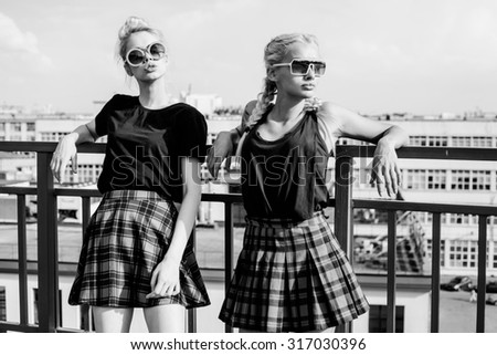 Black white portrait portrait of two pretty hipster blonde sisters  wearing plaid skirt and  black T-shirt. Girls smile, have fun against  urban city.