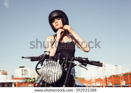 Biker girl sits on vintage custom motorcycle and buttons helmet. Outdoor lifestyle portrait