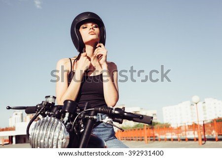 Biker girl sits on vintage custom motorcycle and buttons helmet. Outdoor lifestyle portrait