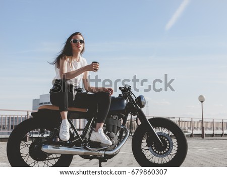 Outdoor lifestyle portrait of sexy biker girl sitting on a vintage custom motorcycle and drinking coffee