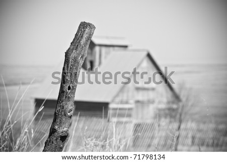 This is a high contrast black and white photo of an old fence post with an old abandoned barn in the background. Shot with a shallow depth of field and slight vignetting.