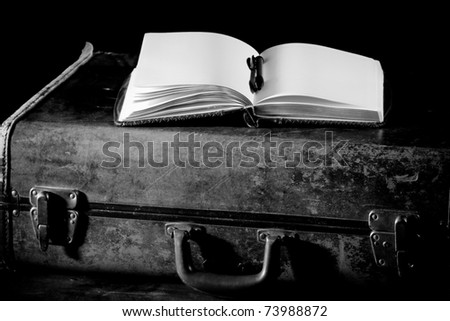 This is a high contrast black and white shot of an old worn down suitcase with an open journal sitting on top.