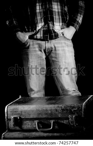 This is a high contrast, black and white image of a young man standing behind a couple old, beat up suitcases with his hands in his pockets.