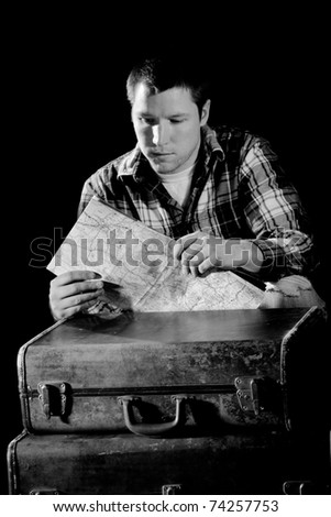 This is a high contrast, black and white image of a young man bent down next to a couple old suitcases looking at a map. Shot with hard light on a black background.