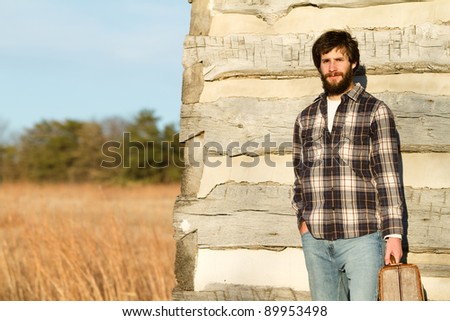 This is a photo of a young man with a beard holding a suitcase leaning against the side of an old cabin.