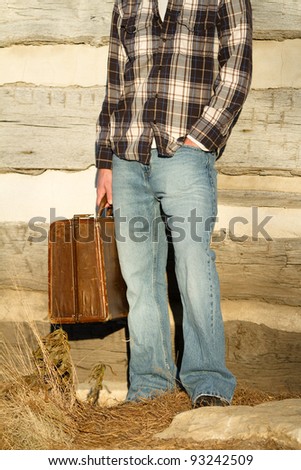 Leaving - This is a photo of a young man leaning against the side of a log cabin holding his old suitcase ready to leave home.