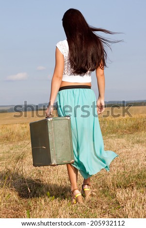 Young girl goes in summer attire comes with a suitcase in hand, goes  on the  field .back view