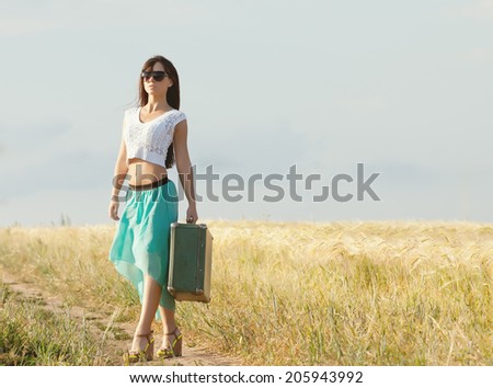 young beautiful long haired girl in glasses and with a suitcase in hand posing on a background field with ears. Front view.  Photo toned light yellow to give summer atmosphere