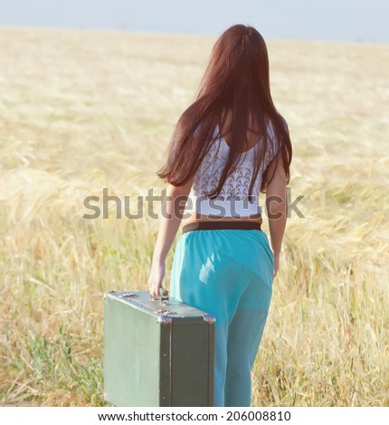 Young girl goes in summer attire comes with a suitcase in hand, goes  on the field .back view