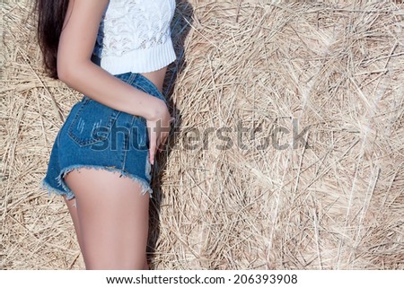 beautiful suntanned female body in short denim shorts and a white blouse on a background of haystack