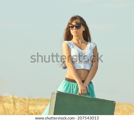 young beautiful long haired girl in glasses and with a suitcase in hand posing on a background field with ears. Front view.
