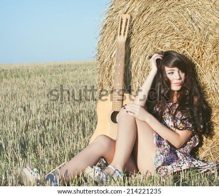 Beautiful country girl in short dress on a summer day basking in the field on a background of haystacks, lies next to a guitar with a bouquet of flowers.Photo tinted light yellow .