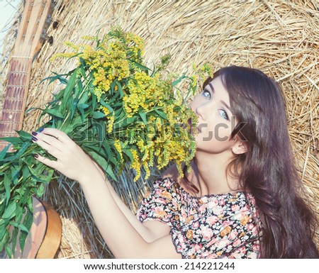 Beautiful young country girl in a short dress posing on a summer day with a bouquet of flowers and a guitar on the background of haystack.Photo tinted light yellow to transfer summer atmosphere