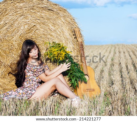 Beautiful young country girl in a short dress posing on a summer day in the field with a bouquet of flowers and a guitar on the background of haystack