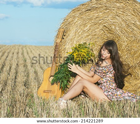 Beautiful young country girl in a short dress posing on a summer day in the field with a bouquet of flowers and a guitar on the background of haystack. Photo tinted light yellow
