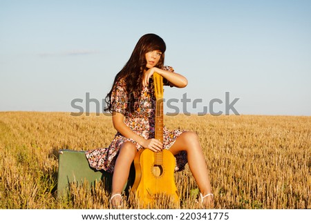 Beautiful young country girl in a short dress posing on a summer day sitting in a field on a suitcase with a guitar.